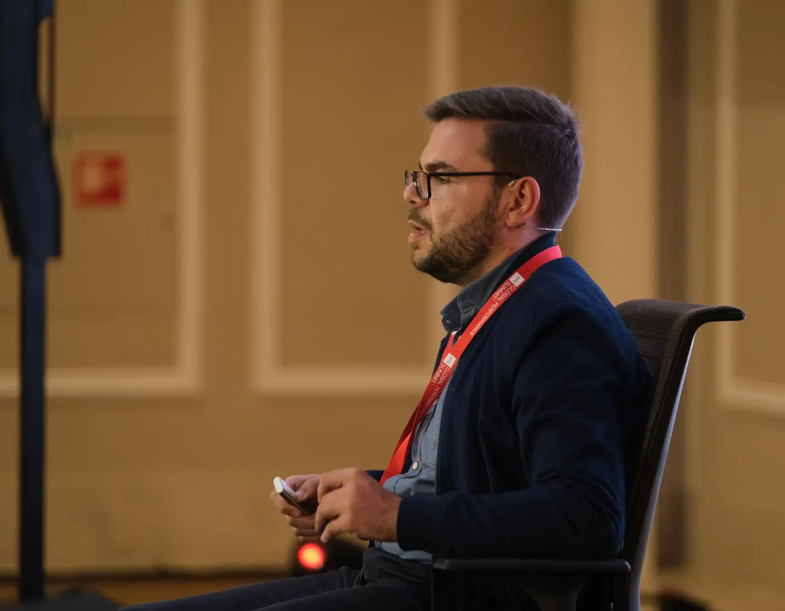 Marco Rodrigues sentado numa cadeira em palco, de microfone de lapela e cartão de orador ao pescoço, a falar para a sala.